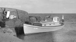 Mustavalkoinen valokuva veneestä kallionreunalla mies nostaa laatikkoa.