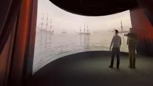 Nuorempi ja vanhempi mieshenkilö katselevat seisaaltaan edessään avautuvaa suurta kangasta, jolle heijastuu vanhoja sota-aluksia merellä, kuvaa kehystävät punaiset esirippuverhot.