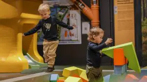 Kaksi leikki-ikäistä poikaa suurien rakennuspalikoiden kimpussa niille tarkoitetulla leikkialuella näyttelytiloissa.