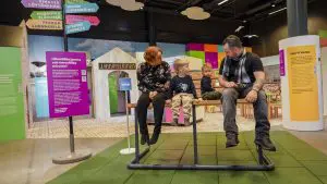 Perhe istuskelee ihmettelemässä tavallista korkeammalla bussipysäkin penkillä näyttelytiloissa.