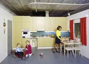 Neuvokas keittiö -julkaisua varten otettu studiokuva. Takaseinällä kellertävät keittiönkaapit. Oikealla seinällä ikkuna jonka vieressä pöytä ja nainen. Kaksi lasta leikkii lattialla.