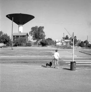Mustavalkoinen kuva. Lapsi ja koira seisovat kävelytiellä suojatin kohdalla. Taustalla kohoaa talo ja Karhulan vesitorni.