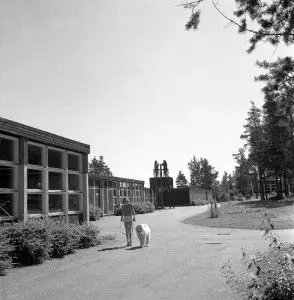 Ruutuikkunainen koulurakennus kuvan vasemmassa laidassa. Tiellä lapsi kävelyttää valkoista koiraa.