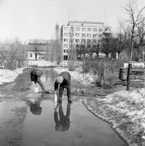 Kuvassa on puisto. Lunta ja loskaa. Kaksi lasta leikkii vesilätäkössä. Taustalla kerrostalo.