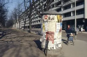 Katunäkymää reunystaa lehdettömät puut ja valkoinen kerrostalo. Etualalla mainospylväs ja nainen joka työntää lastenrattaita.