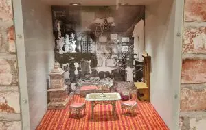 vitriinin taustalla kuva hienosta asunnosta. Vitriinissä metalliset, hienot nukkekodin salin kalusteet. Punainen matto.