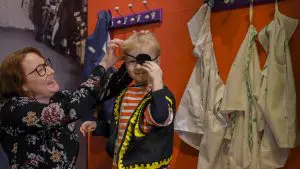 Pieni hymyilevä merirosvoksi pukeutunut nuoriherra katsoo kameraan isoäidin auttaessa pukeutumisessa.