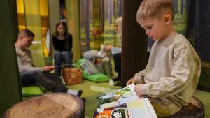 Etualalla keskittyneestä lastenkirjaa lukeva pikkupoika ja taustalla perheen äiti, isä ja teinit istuskelevat leikkimetsässä.