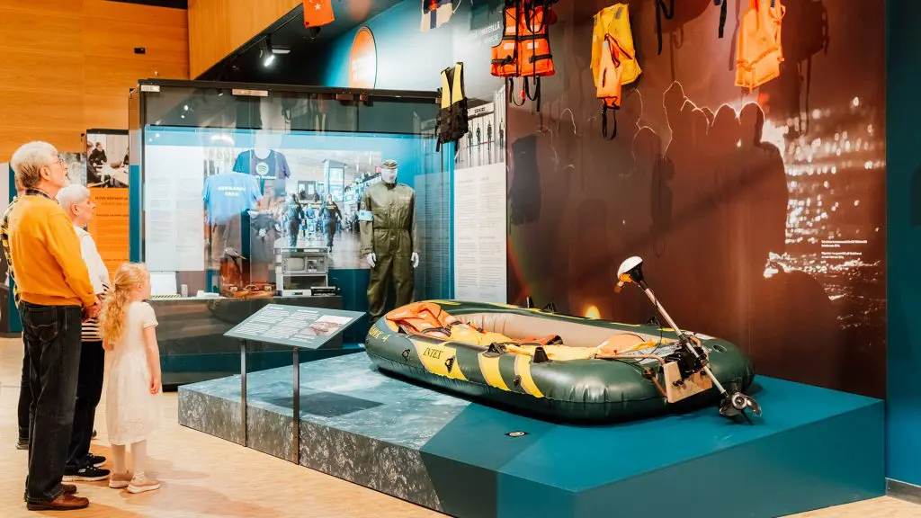 Perhe kolmessa sukupolvessa katselee kumista pelastusvenettä ja seinällä roikkuvia pelastusliivejä.