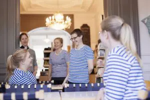 Neljä iloista naista sinivalkoraidallisissa paidoissa pöytien äärelle kokoontuneina, taaempana seisovalla naisella harmaan neuleen alla papin kaulus.