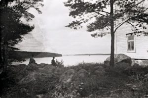 Mustavalkoisessa vanhassa valokuvassa kaksi ihmistä istuvat eri kivillä ja katselevat vesistön suuntaan, kuvan oikealla laidalla puutalon kulma.