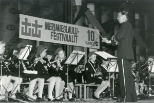 Nuori puhallinorkesteri soittaa nuoteista kapelimestarin seisoessa etualalla, yläpuolella Merimieslaulufestivaalien 10-vuotisjuhlakyltti.
