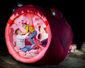 Äiti pitelee lasta Merimonterit-näyttelystä löytyvän merimakkaran suulla, sivusta kurkistaa pieni tyttö