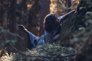 Tanssitaiteilija Aino Karstinen hämyisessä metsässä puuhun nojaten