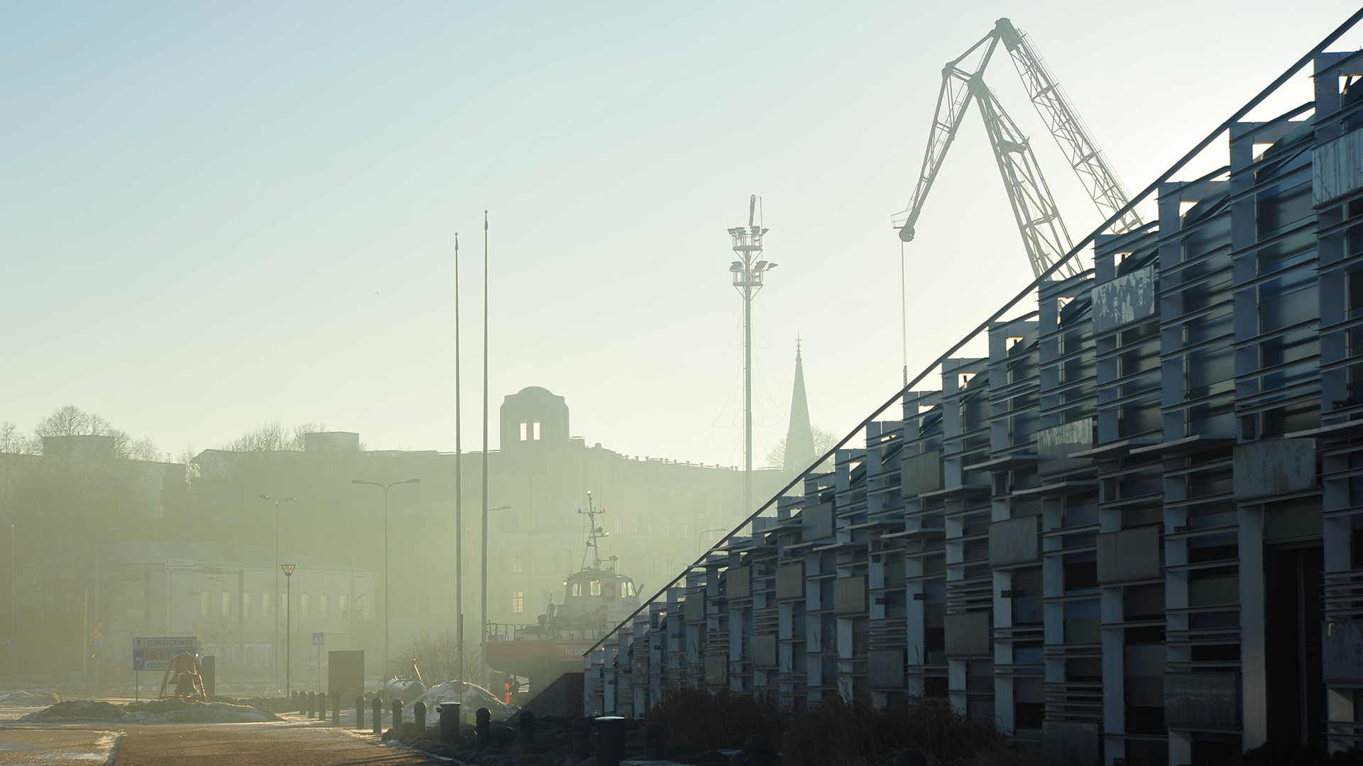 Kuvan oikean reunan peittää diagonaalisesti Vellamon rakennus, jonka takaa pilkistää satamanosturi. Taustamaisema jää usvaiseksi.