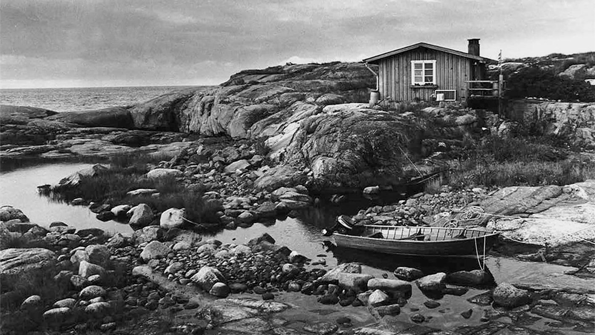 Mustavalkoinen valokuva saaristosta; kalliolla pieni mökki ja etualalla puinen vene.