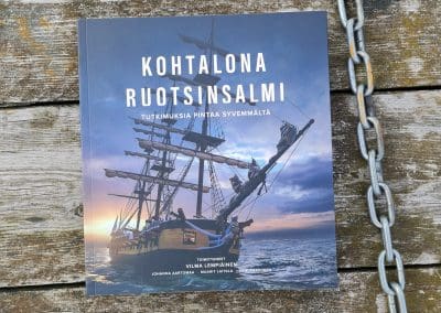 Kohtalona Ruotsinsalmi -kirja kuvattuna rouheaa laudoitusta vasten, kirjan vieressä kulkee kettinki.